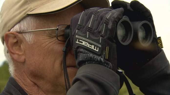 A man wearing a beige cap and dark-coloured gloves uses binoculars.
