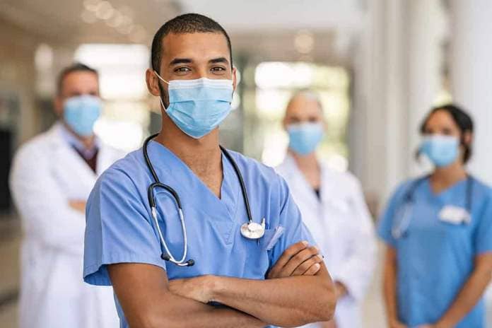 Confident multiethnic male nurse in front of his medical team looking at camera wearing face mask during covid-19 outbreak. Happy and proud indian young surgeon standing in front of his colleagues.