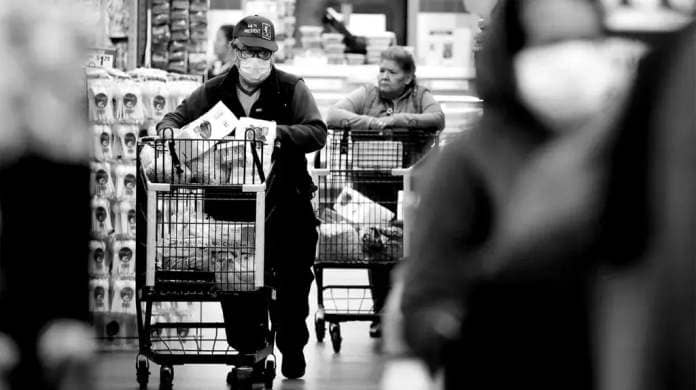 A scene from a supermarket as an older male adult pushes his shopping cart full of items as female shopper does the same in the background