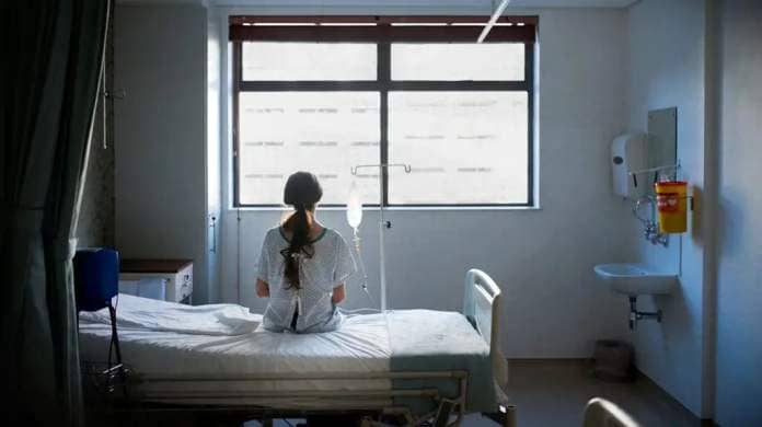 A young woman gazes out a window while sitting on a bed in a hospital