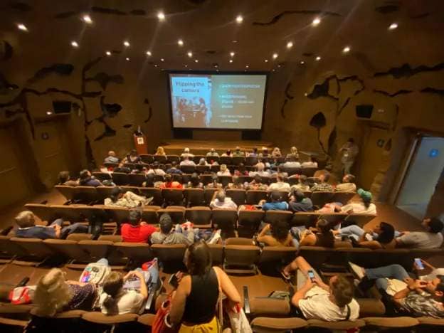 A woman leads a lecture inside of a modern auditorium. There is a slide projected on a screen up front and the auditorium is filled with rows of people.
