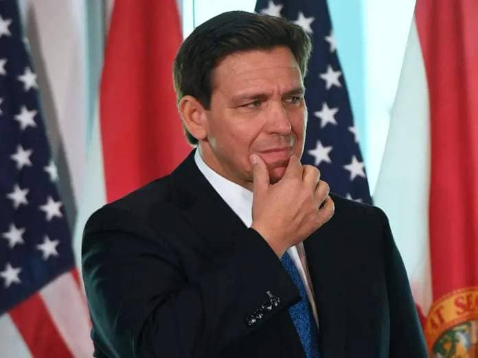 Ron DeSantis stroking his chin while wearing a black suit with a blue tie in a large room with American flags and Florida flags behind him.