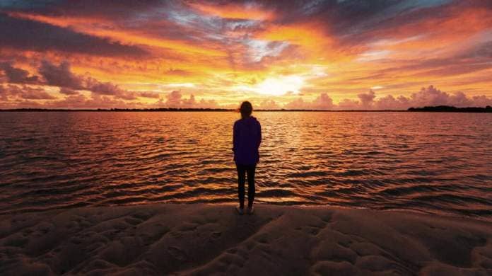 A woman looks out into the sunset with ehr back turned against the camera in front of a body of water