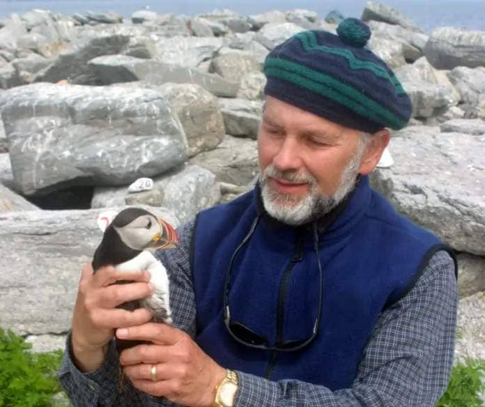 A man wearing a blue vest, a plaid, long-sleeved shirt and a striped paddy cap looks at an Atlantic puffin that he's holding.