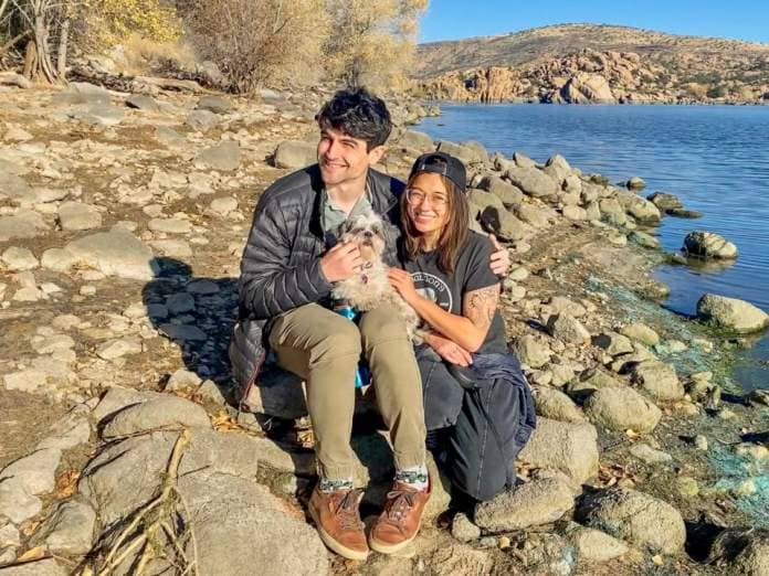 A dark-haired couple holding a small, white dog sits on rocky, pebbled shore with a lake on the right and hills and clear blue skies in the background.