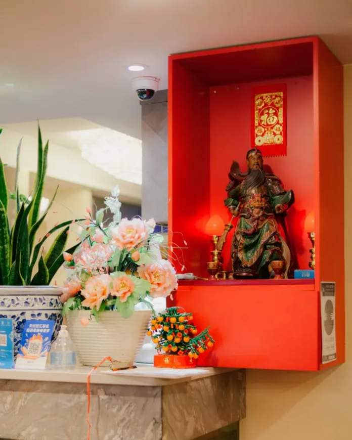A Chinese deity in a red case on a wall at Golden Paramount, with a vase filled with blousy orange-pink blooms and a houseplant on a marble shelf in front of it