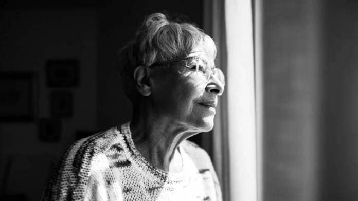 An older woman of color with gray hair and wearing glasses looks outside the window from a dark room