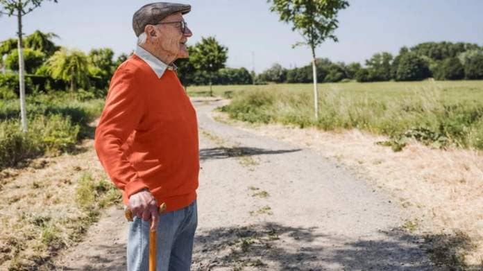 An older man wearing a hat and holding a cane looks into the distance while going on a walk outside