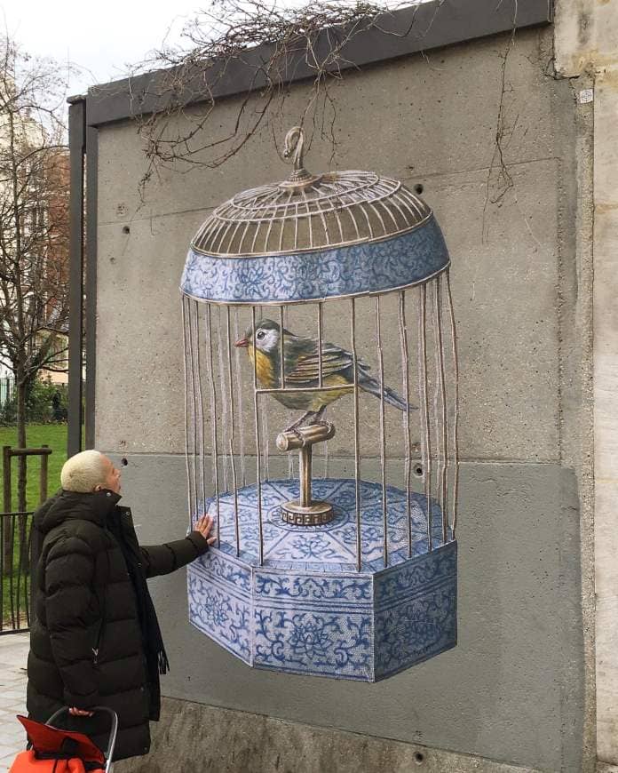 a woman in a puffy black coat touches the ornate blue and white cage of a yellow bird on a perch in a wheatpaste