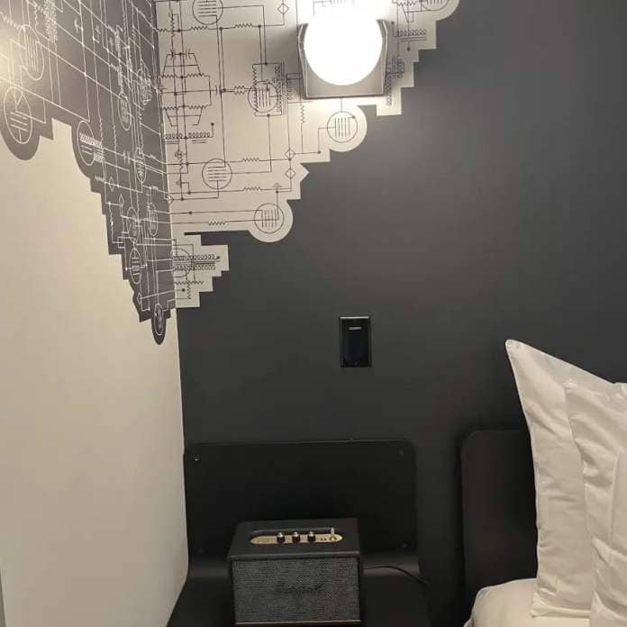Compact hotel room with schematic wall art, a bedside table featuring a Marshall speaker, and minimalistic furnishings