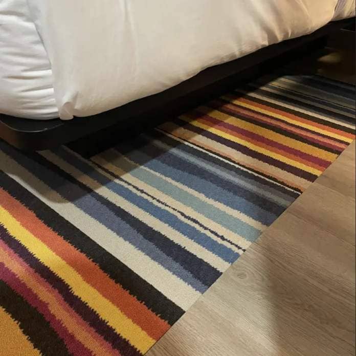 A multicolored striped rug extends from underneath a bed onto a wooden floor in a hotel room setting
