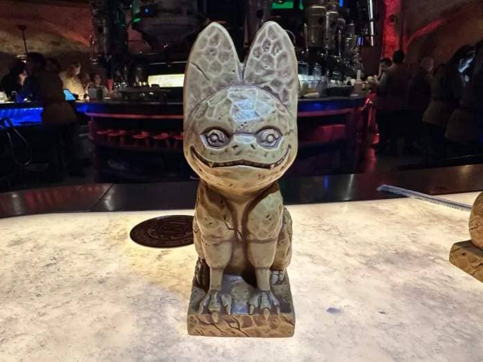 Statue of a stylized alien creature at a themed bar with ambient lighting and patrons in the background.