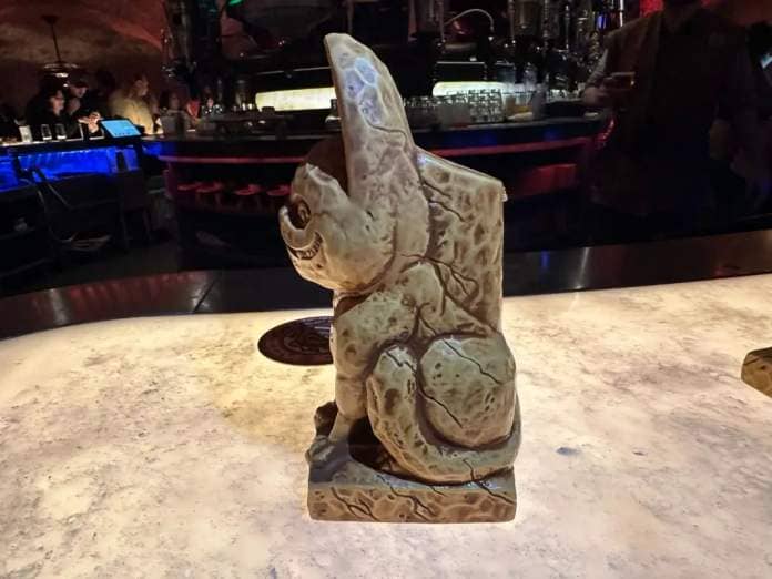 A carved stone elephant sculpture displayed on a bar counter, with dim lighting and indistinct figures in the background.